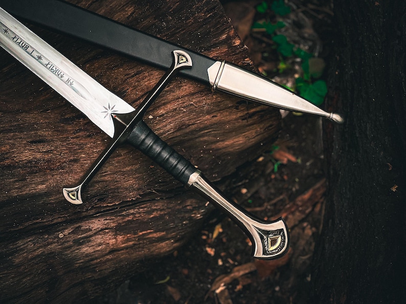 May include: A detailed image of a sword and scabbard resting on a wooden surface. The sword features a silver blade with etched text and a star design. The handle is wrapped in black leather, and the scabbard is black with silver accents.