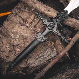 May include: A black fantasy sword with an ornate hilt featuring a skull design and a white blade. The sword rests on a pile of weathered wood logs and branches, creating a rustic and dramatic scene.