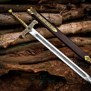 May include: A decorative sword with a silver blade and ornate gold hilt, featuring a blue gem. The sword rests on a bed of wooden logs, with a brown sheath adorned with gold accents. The sword's design suggests a medieval or fantasy theme.