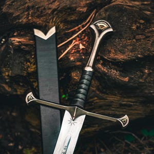May include: A detailed image of a sword with a silver blade and a black leather-wrapped handle. The hilt and crossguard are silver with gold accents. The sword rests against a black sheath, set against a dark, textured background.