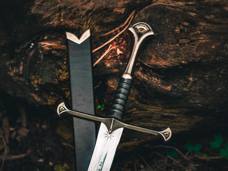 May include: A detailed image of a sword with a silver blade and a black leather-wrapped handle. The hilt and crossguard are silver with gold accents. The sword rests against a black sheath, set against a dark, textured background.
