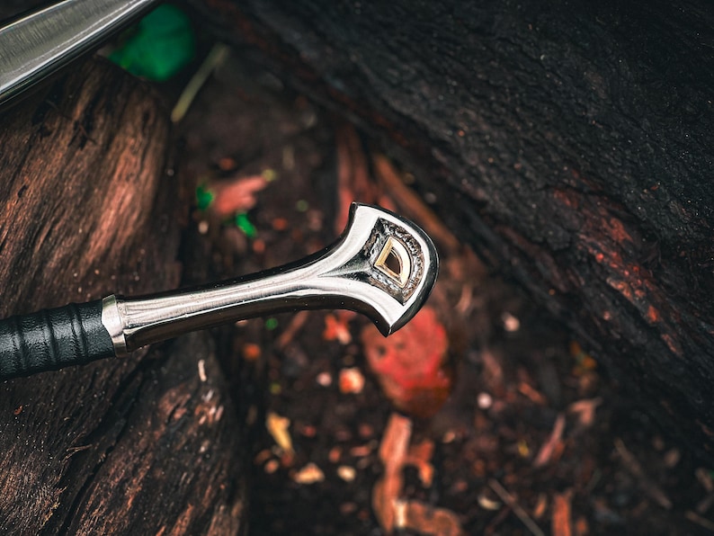 May include: Close-up of a sword handle with a silver finish and a decorative gold and silver emblem. The handle is wrapped in black leather. The sword rests on a bed of dark wood and foliage.