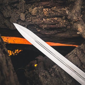 May include: A silver sword blade with a pointed tip, set against a backdrop of dark, textured wood and a fiery orange glow. The blade has three parallel grooves running down its length, reflecting the light.