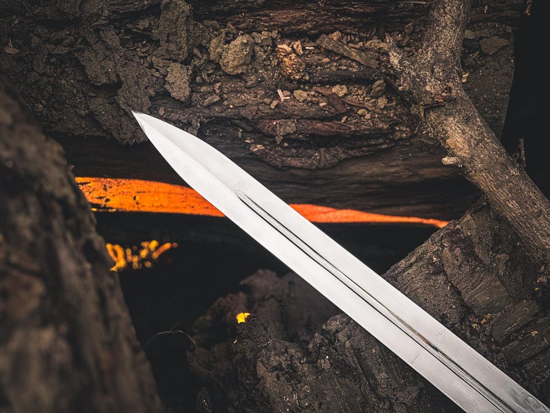 May include: A silver sword blade with a pointed tip, set against a backdrop of dark, textured wood and a fiery orange glow. The blade has three parallel grooves running down its length, reflecting the light.