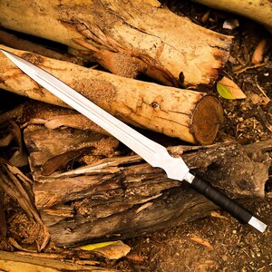 Peut inclure: Une &eacute;p&eacute;e argent&eacute;e avec une poign&eacute;e noire repose sur une pile de b&ucirc;ches en bois. La lame est longue et tranchante, avec une pointe ac&eacute;r&eacute;e. La poign&eacute;e est envelopp&eacute;e d'un mat&eacute;riau noir. Les b&ucirc;ches sont de diff&eacute;rentes nuances de brun.