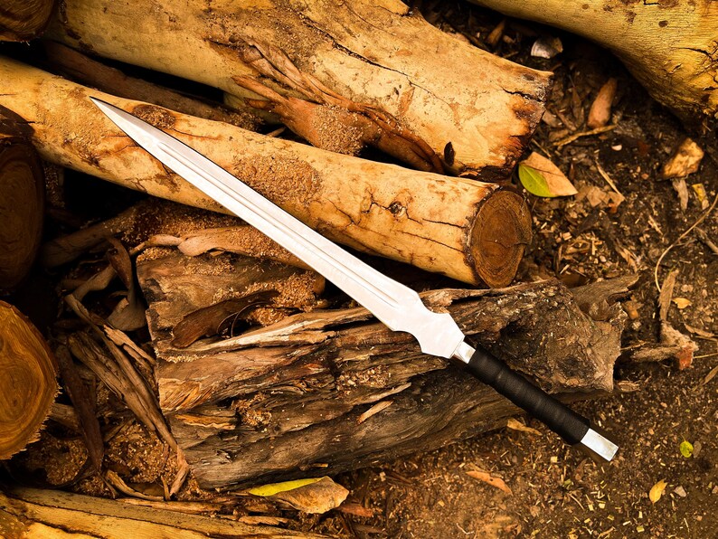 Peut inclure: Une &eacute;p&eacute;e argent&eacute;e avec une poign&eacute;e noire repose sur une pile de b&ucirc;ches en bois. La lame est longue et tranchante, avec une pointe ac&eacute;r&eacute;e. La poign&eacute;e est envelopp&eacute;e d'un mat&eacute;riau noir. Les b&ucirc;ches sont de diff&eacute;rentes nuances de brun.
