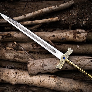 May include: A decorative sword with a silver blade and a gold-colored hilt. The hilt features an ornate design with a blue gem. The sword is lying on a pile of wooden logs, creating a rustic aesthetic.