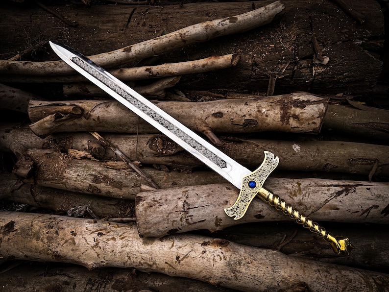 May include: A decorative sword with a silver blade and a gold-colored hilt. The hilt features an ornate design with a blue gem. The sword is lying on a pile of wooden logs, creating a rustic aesthetic.