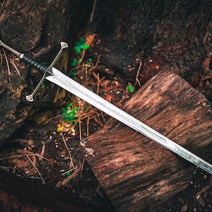 May include: A silver sword with a black handle and ornate hilt rests on a weathered wooden surface. The blade has etched inscriptions. The sword is a fantasy-themed item, likely for display or cosplay.