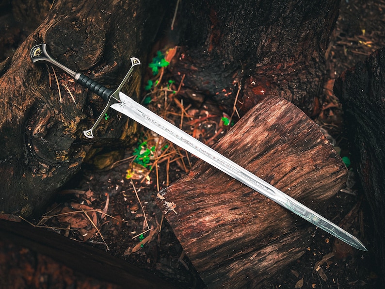 May include: A silver sword with a black handle and ornate hilt rests on a weathered wooden surface. The blade has etched inscriptions. The sword is a fantasy-themed item, likely for display or cosplay.
