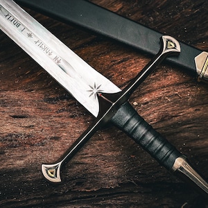 May include: A close-up of a medieval sword with a silver blade and a black leather-wrapped handle. The sword features a crossguard with gold accents and is resting on a wooden surface. The scabbard is black with gold details.