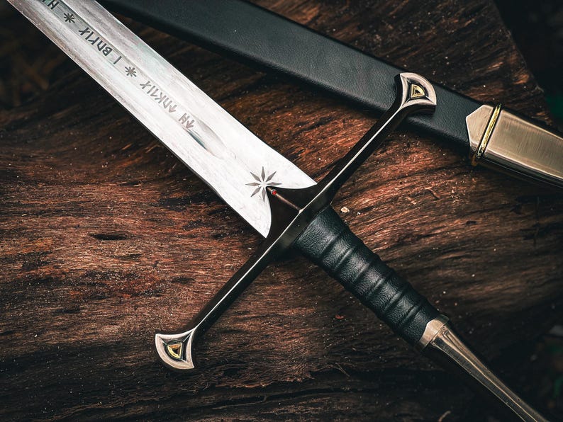 May include: A close-up of a medieval sword with a silver blade and a black leather-wrapped handle. The sword features a crossguard with gold accents and is resting on a wooden surface. The scabbard is black with gold details.