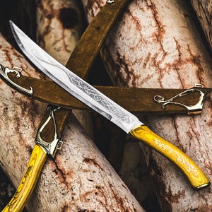 May include: A decorative knife with a silver blade and a yellow handle. The handle has a curved design with gold accents. The knife is resting on a brown leather sheath, with a wooden background. The knife's blade has an ornate pattern.