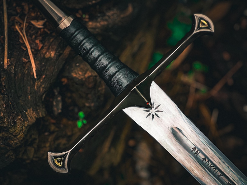 May include: A detailed image of a sword with a silver blade and black handle. The blade features a starburst design and etched markings. The hilt has gold accents. The sword is set against a blurred, natural background.