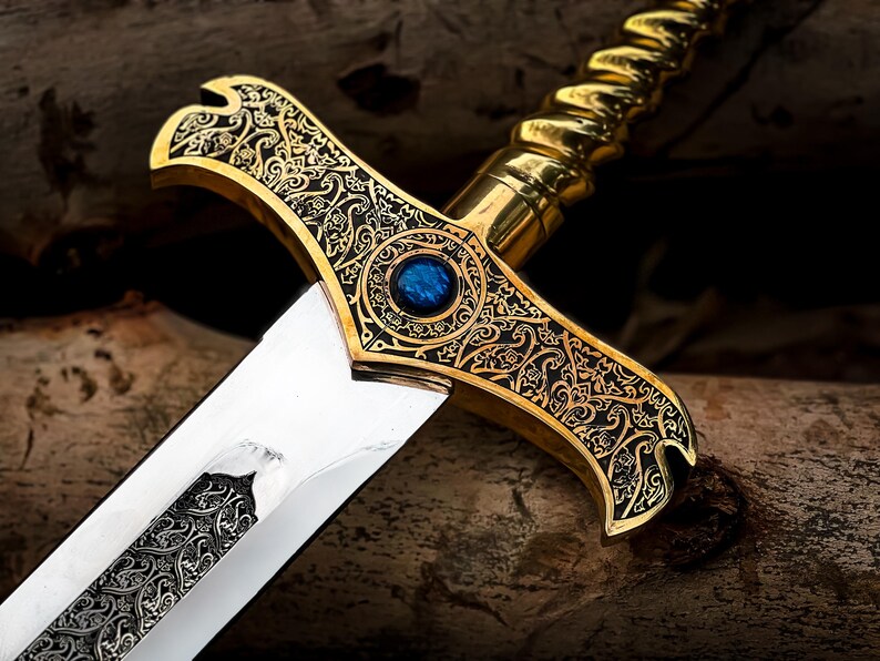 May include: Close-up of an ornate sword with a gold-colored hilt and a silver blade. The hilt features intricate black and gold filigree designs and a blue gemstone. The blade has a detailed pattern. The sword is a decorative item.