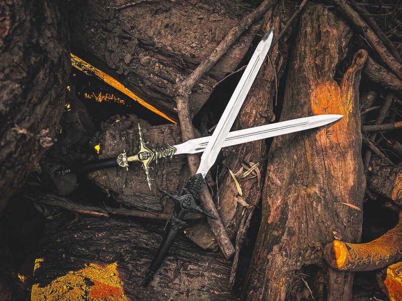 May include: Two crossed swords with silver blades and ornate handles rest on a bed of wood and branches. One sword has a black handle and a detailed, dark metal crossguard. The other has a gold and silver handle.