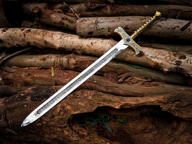 May include: A decorative sword with a silver blade and a gold-colored hilt. The hilt features intricate designs and a blue gem. The sword is resting on a pile of wooden logs, creating a rustic and fantasy-themed aesthetic.