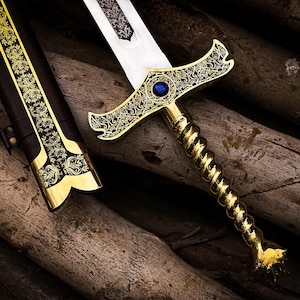May include: An ornate sword with a gold-colored hilt and a blue gem set in the crossguard. The blade is silver, and the handle has a twisted design. The scabbard is black with gold accents. The sword is resting on a bed of wooden logs.