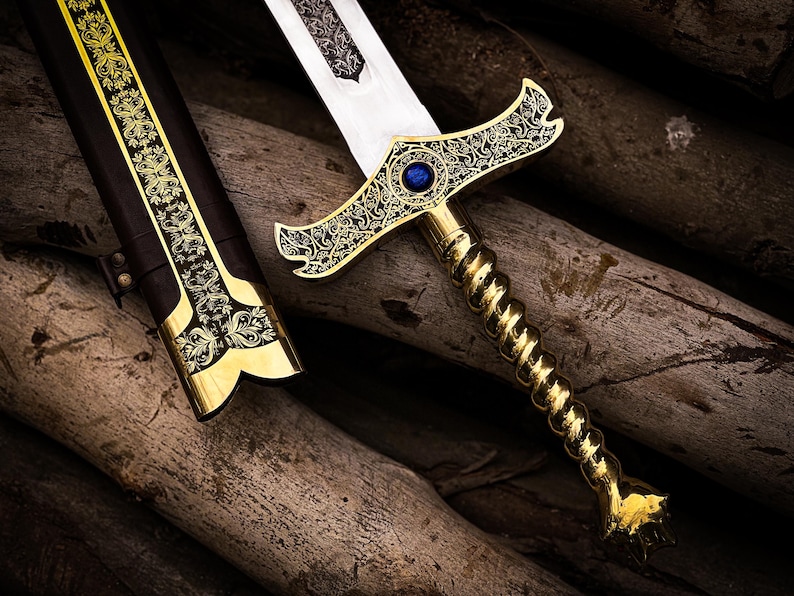 May include: An ornate sword with a gold-colored hilt and a blue gem set in the crossguard. The blade is silver, and the handle has a twisted design. The scabbard is black with gold accents. The sword is resting on a bed of wooden logs.