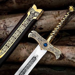 May include: An ornate sword with a gold-colored handle and crossguard, featuring intricate engraved patterns. The blade is silver, and a blue gem is set in the center of the crossguard. A matching scabbard with gold accents lies beside it.