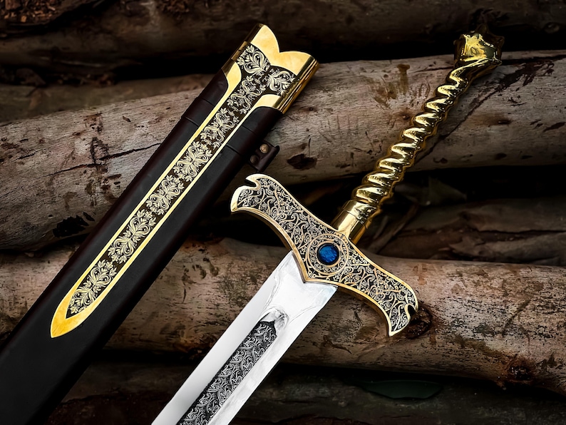 May include: An ornate sword with a gold-colored handle and crossguard, featuring intricate engraved patterns. The blade is silver, and a blue gem is set in the center of the crossguard. A matching scabbard with gold accents lies beside it.
