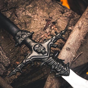 May include: A black fantasy sword with a skull design on the hilt and crossguard, resting on a wooden surface. The blade is silver. The sword is a decorative item.
