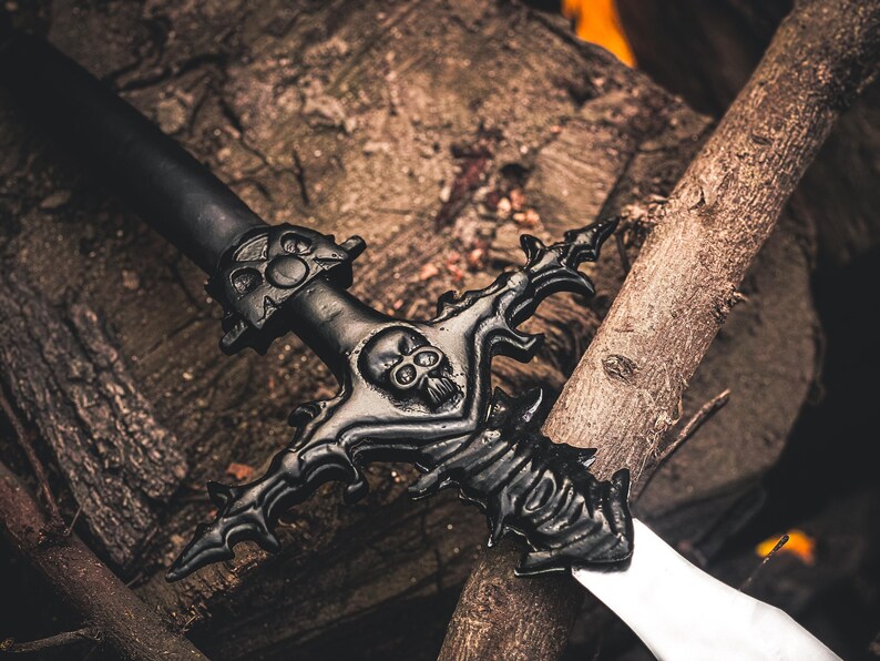 May include: A black fantasy sword with a skull design on the hilt and crossguard, resting on a wooden surface. The blade is silver. The sword is a decorative item.