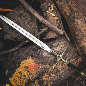 May include: A silver sword with an ornate gold and black handle rests on a bed of weathered wood and fallen branches. The blade is long and sharp, reflecting the light. The hilt features intricate designs, suggesting a historical or fantasy theme.