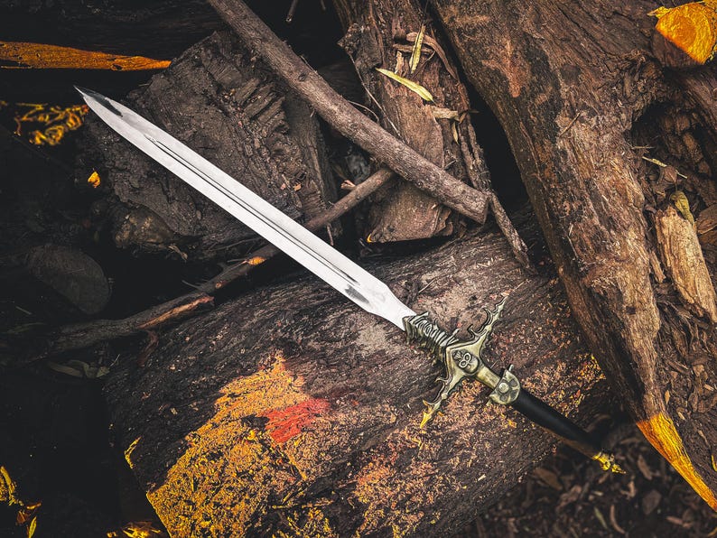 May include: A silver sword with an ornate gold and black handle rests on a bed of weathered wood and fallen branches. The blade is long and sharp, reflecting the light. The hilt features intricate designs, suggesting a historical or fantasy theme.