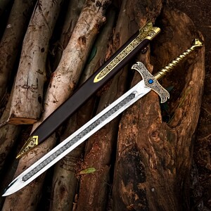 May include: A decorative sword with a silver blade and ornate gold hilt, resting on a wooden surface. The sword has a detailed design and a blue gem. A brown sheath with gold accents lies beside it.