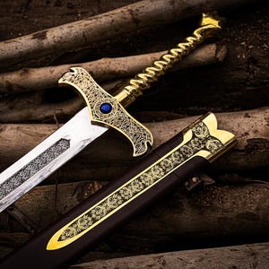 May include: A detailed, ornate sword with a gold-colored hilt and a silver blade. The hilt features intricate designs and a blue gem. The sword rests beside a brown sheath with gold accents, all set against a backdrop of wooden logs.