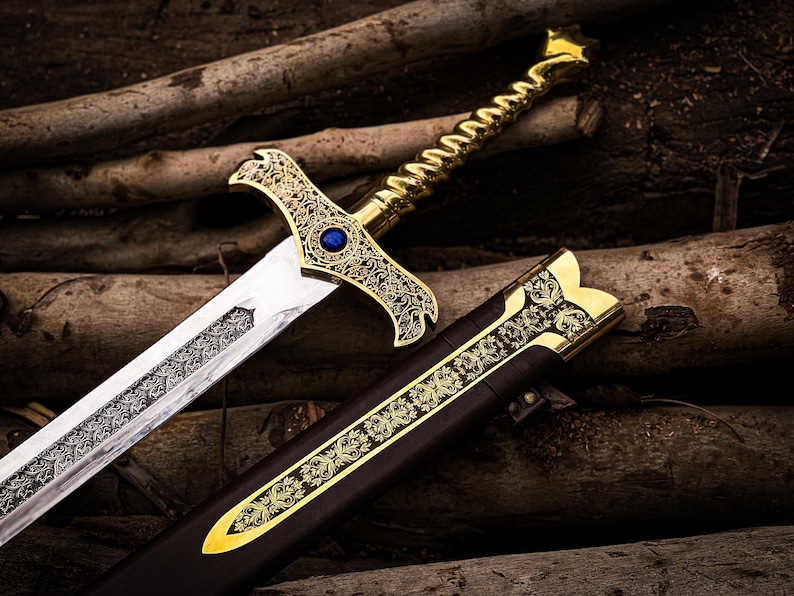 May include: A detailed, ornate sword with a gold-colored hilt and a silver blade. The hilt features intricate designs and a blue gem. The sword rests beside a brown sheath with gold accents, all set against a backdrop of wooden logs.