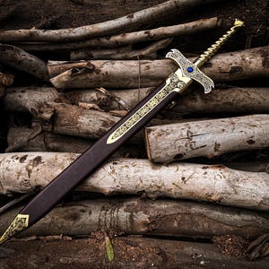 May include: A decorative sword with a gold-colored hilt and accents, a blue gem, and a brown sheath. The sword is resting on a pile of logs, creating a rustic and fantasy-themed image.