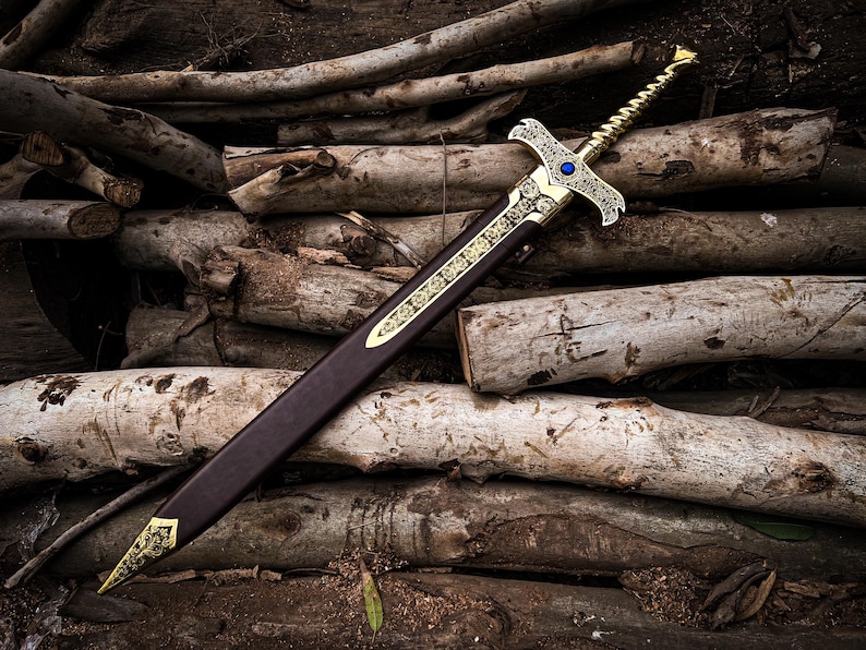 May include: A decorative sword with a gold-colored hilt and accents, a blue gem, and a brown sheath. The sword is resting on a pile of logs, creating a rustic and fantasy-themed image.