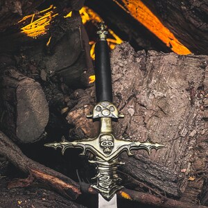 May include: A detailed image of a decorative sword with a black handle and a silver blade. The sword features an ornate gold-colored hilt with a skull design. The sword is set against a backdrop of stacked logs and glowing embers.