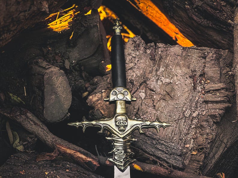 May include: A detailed image of a decorative sword with a black handle and a silver blade. The sword features an ornate gold-colored hilt with a skull design. The sword is set against a backdrop of stacked logs and glowing embers.