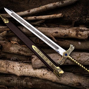 May include: A detailed image of a decorative sword with a silver blade and gold hilt. The hilt features an ornate design with a blue gem. The sword rests beside a brown sheath with gold accents, all set against a backdrop of wooden logs.