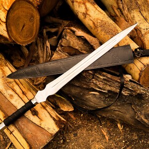 Peut inclure: Une &eacute;p&eacute;e argent&eacute;e avec une poign&eacute;e noire et un fourreau en cuir est pos&eacute;e sur une pile de b&ucirc;ches. L'&eacute;p&eacute;e a une longue lame pointue et une garde d&eacute;corative. Le fourreau est noir et poss&egrave;de une sangle.