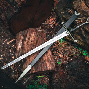 May include: A silver sword with a black handle and crossguard, resting on a black sheath. The sword is displayed on a bed of wood logs and forest debris, creating a rustic aesthetic.