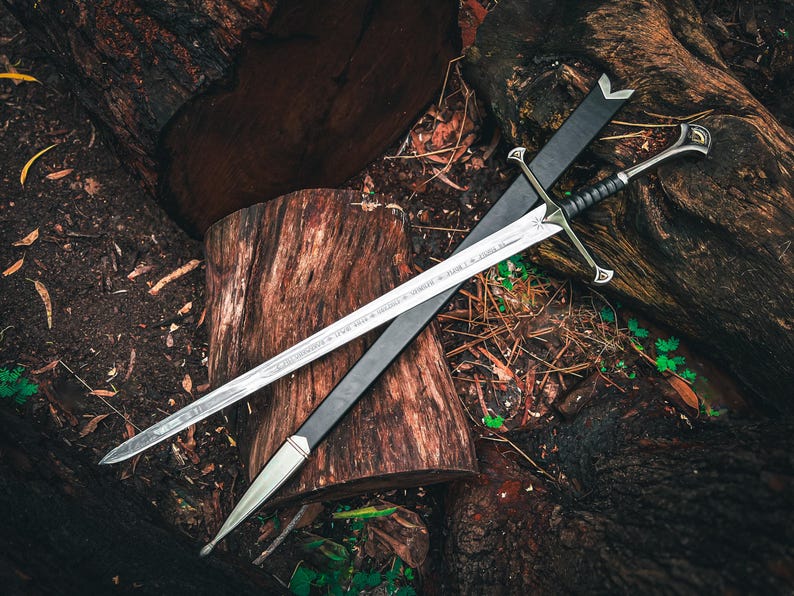 May include: A silver sword with a black handle and crossguard, resting on a black sheath. The sword is displayed on a bed of wood logs and forest debris, creating a rustic aesthetic.