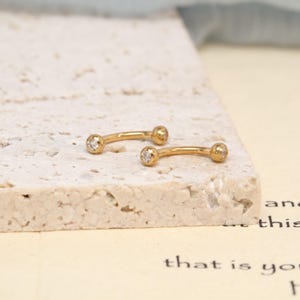 K&ouml;nnte beinhalten: Zwei goldfarbene, gebogene Barbell-K&ouml;rperschmuckst&uuml;cke, die jeweils an einem der abgerundeten Enden mit einem kleinen, klaren, rund geschliffenen Edelstein besetzt sind. Der Schmuck hat eine polierte metallische Oberfl&auml;che und eine gebogene Form.