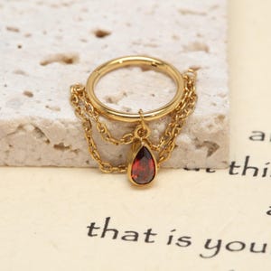 May include: A gold-toned hoop ring featuring two delicate dangling chains and a single teardrop-shaped red gemstone charm. The metal ring has a smooth finish and is designed with a hinged closure mechanism for secure placement.