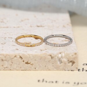 May include: Two circular hoop rings, one in a gold tone and one in a silver tone, each featuring a row of small, clear, sparkling stones embedded along the outer edge. Both rings have a hinged closure mechanism visible on the side.