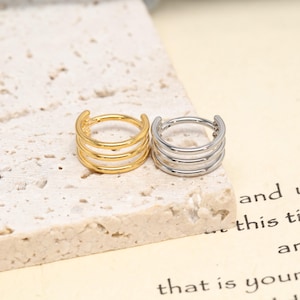 May include: Two triple-band hoop earrings, one in a polished gold tone and one in a polished silver tone. Each earring features three parallel circular bands connected at a single point, creating a layered, stacked appearance for the ear.