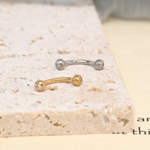 K&ouml;nnte beinhalten: Zwei gebogene Piercing-St&auml;be, einer in Silberoptik und einer in Goldoptik. Jeder Stab verf&uuml;gt &uuml;ber einen kleinen, klaren, runden Schmuckstein, der in die Kugel an jedem Ende des gebogenen Metallstabs eingelassen ist, was ein schlichtes Design ergibt.