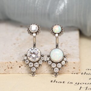 May include: Two navel rings featuring a curved barbell design. The left ring has a clear round stone at the top and a larger clear stone at the bottom with small accent stones. The right ring has an iridescent opal stone at the top and bottom.