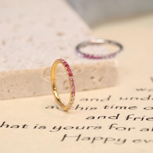 Op de afbeelding: Twee ringen worden getoond, één goudkleurig en één zilverkleurig. De gouden ring heeft een verloop van roze edelstenen, van donker naar licht. De zilveren ring is bezet met roze edelstenen. De ringen staan tegen een neutrale achtergrond.