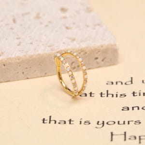 May include: A gold-coloured double hoop earring. One hoop is decorated with small, clear gemstones, while the other features small, white gemstones. The earring is set against a neutral background.