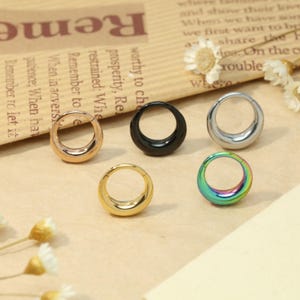 May include: A collection of six hoop earrings in various colors, including gold, silver, black, and iridescent. The earrings are circular with a curved, teardrop-like design. They are displayed on a light brown surface.