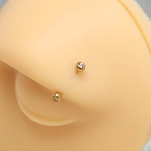 K&ouml;nnte beinhalten: Ein goldener, gebogener Barbell-Piercingschmuck mit zwei runden Enden, die jeweils mit einem kleinen, klaren, kreisf&ouml;rmigen Edelstein besetzt sind. Der Schmuck liegt auf einer glatten, hellen Oberfl&auml;che und zeigt das metallische Finish.